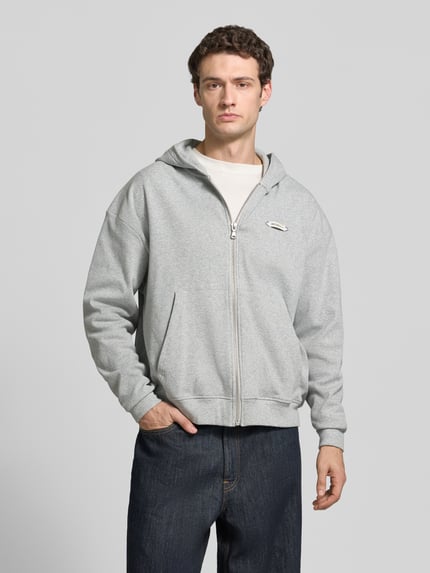 Männermodel trägt graue Regular Fit Sweatjacke mit Känguru-Tasche Modell 'L’ATELIER LINE' von RETERNITY, Label-Detail, Ganzkörperansicht von vorne. Baumwoll-Elasthan-Mix, Kapuze, Reißverschluss, Rückenlänge und Ärmellänge gemäß Größe M.