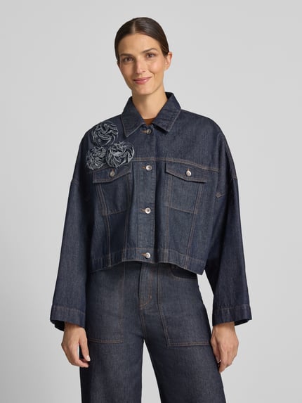 Vrouwenmodel draagt Cropped Jeansjacke aus reiner Baumwolle van DRYKORN in donkerblauw met bloemenapplicatie, puur katoen, los model, platte kraag, doorlopende knoopsluiting, borstzakken, cropped, borstwijdte 72 cm, ruglengte 51 cm, schouderbreedte 43 cm, mouwlengte 60 cm, vooraanzicht van het volledige lichaam.