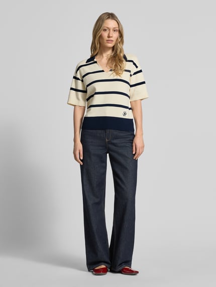 Frauenmodel trägt Regular Fit Pullover aus reiner Baumwolle von Tommy Hilfiger mit Allover-Streifenmuster, Polokragen, V-Ausschnitt, Logo-Stitching, kurzarm, Feinstrick, in Creme und Dunkelblau, kombiniert mit dunkelblauer Jeans und roten Schuhen, Ganzkörperansicht von vorne.