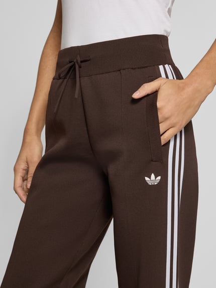 Frauenmodel trägt Regular Fit Sweathose mit Logo-Detail von Adidas Originals, uni braun, weiße Streifen, Zugband, seitliche Taschen, Nahaufnahme Oberkörper.