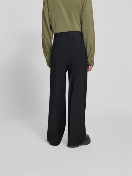 Männermodel trägt Wide Fit Stoffhose aus Woll-Mix von Filippa K in Schwarz mit Bügelfalten und Gesäßtaschen, Ganzkörperansicht Rückansicht.