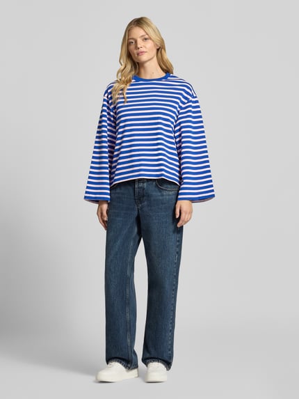 Frauenmodel trägt Longsleeve im Allover-Look mit Rundhalsausschnitt von Gina Tricot, blau-weiß gestreift, Relaxed Fit, reine Baumwolle, kombiniert mit Jeans und weißen Sneakern, Ganzkörperansicht von vorne.