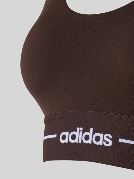 Vrouwenmodel draagt 'BH met elastische logoband' van adidas Sportswear in donkerbruin, stretchmateriaal, detailopname bovenlichaam.