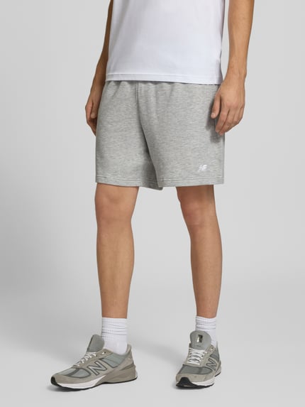 Männermodel trägt graue Sweatshorts aus Baumwoll-Mix Modell 'FRENCH' von New Balance, elastischer Bund, Logo, Nahaufnahme Beinbereich, Ganzkörperansicht von vorne.