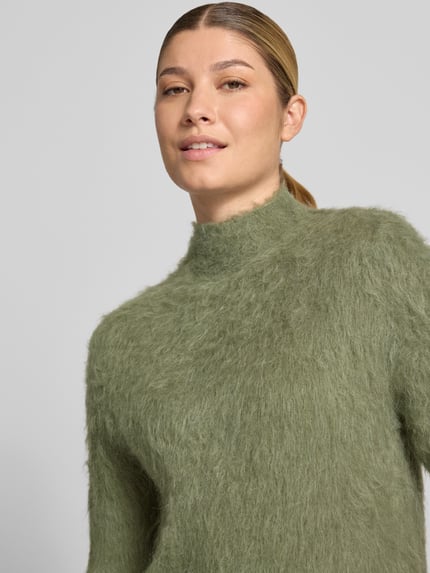 Frauenmodel trägt Strickshirt aus Alpaka-Mix mit kurzem Stehkragen von someday in Olivgrün, flauschige Textur, Nahaufnahme Oberkörper. Körpernaher Schnitt, Modell Tuomina, 1/2-Arm, Rückenlänge 60 cm, Ärmellänge 44 cm.