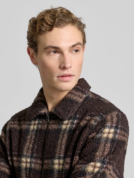 Männermodel trägt Hemdjacke mit Reißverschluss von Forét aus braunem, kariertem Teddyfell mit Kentkragen, Nahaufnahme Oberkörper.