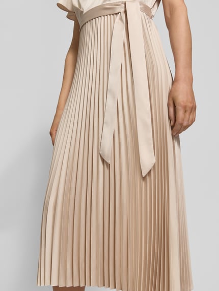 Frauenmodel trägt Midikleid mit Plisseefalten und V-Ausschnitt von comma, beigefarbener Stoff mit Bindegürtel, Nahaufnahme Oberkörper bis Knie. Material-Mix mit Stretch-Anteil und tailliertem Schnitt.