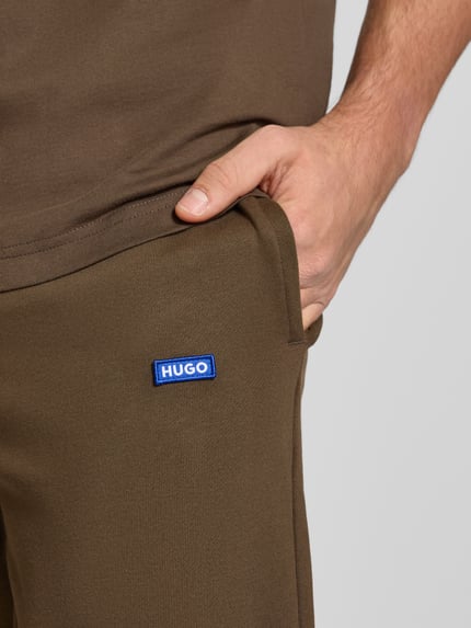Mężczyznamodel nosi Comfort Fit Sweatpants aus reiner Baumwolle Modell 'NAPIN' od Hugo Blue w jednolitym brązowym kolorze z naszywką z logo, kieszenie boczne, elastyczny pas, materiał czysta bawełna, detalami w zbliżeniu na górną część ciała.