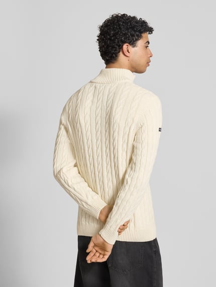 Männermodel trägt Strickpullover aus reiner Baumwolle von Superdry mit Zopfmuster, Troyer-Kragen, Label-Detail, Gerippte Abschlüsse, Rückenlänge 70 cm, Ärmellänge 71 cm, enge Passform, cremefarben, Ganzkörperansicht Rückansicht.