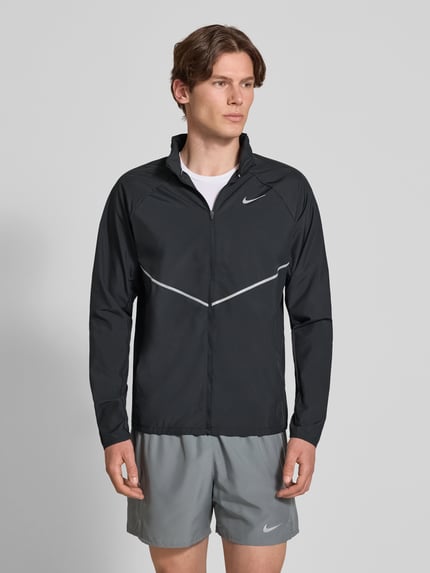 Männermodel trägt Regular Fit Jacke mit Stehkragen Modell 'Miler' von NIKE TRAINING in Schwarz mit Logo-Print und Reißverschluss, Ganzkörperansicht von vorne. Rückenlänge bei Größe M: 67 cm, Ärmellänge bei Größe M: 64 cm.