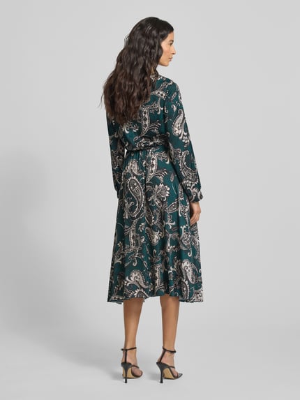 Frauenmodel trägt Cocktailkleid mit Rundhalsausschnitt von Rinascimento im Material-Mix mit Stretch-Anteil, körpernahe Passform, Plisseefalten, farbiges Muster in Grün und Beige, Rückenlänge 113 cm, Ganzkörperansicht Rückansicht.