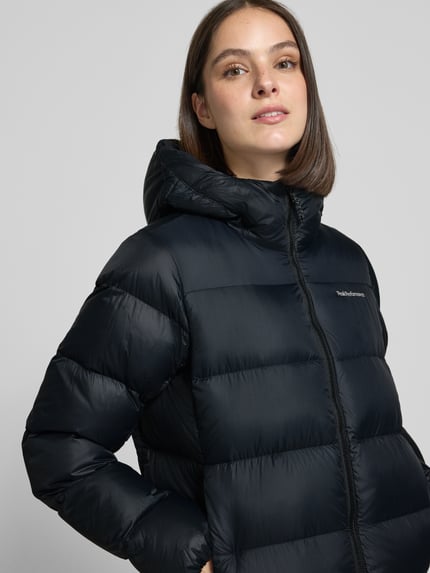 Frauenmodel trägt Steppmantel mit Daunen-Federn-Füllung Modell 'FROST' von Peak Performance in Schwarz, glatte Textur, Nahaufnahme Oberkörper.