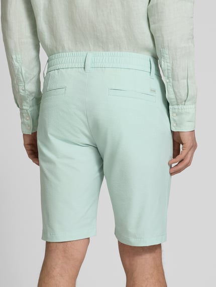 Männermodel trägt Tapered Fit Bermuda mit Kordelzug Modell 'CHINO' von BOSS Orange in Mint, Allover-Muster, Crinkle-Textur, Ganzkörperansicht Rückansicht.