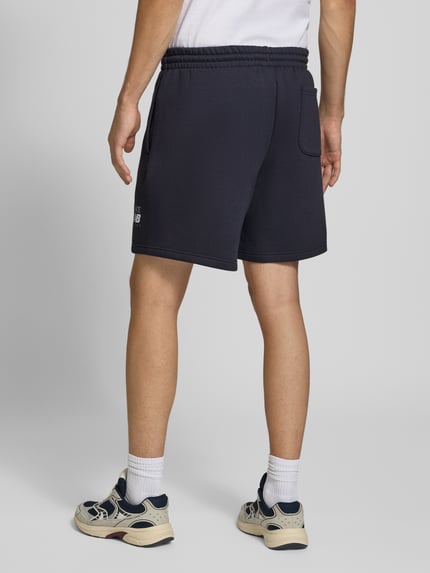Männermodel trägt dunkelblaue Sweatshorts aus Baumwoll-Mix von New Balance mit elastischem Bund, Label-Print, Gesäßtasche, gerade Passform, Ganzkörperansicht Rückansicht.
