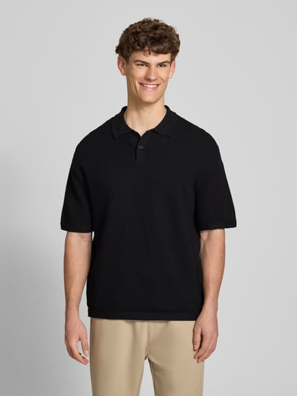 Männermodel trägt Relaxed Fit Poloshirt Modell 'FERDINAND' von Redefined Rebel aus Reine Baumwolle, Strickshirt, Knopfleiste, Rückenlänge 70 cm bei Größe M, schwarze Struktur, beige Hose, Ganzkörperansicht von vorne.