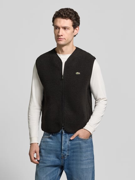 Männermodel trägt Weste mit Fleece-Struktur von Lacoste in Schwarz, unifarben mit Logo-Patch, Reißverschlusstaschen, Ganzkörperansicht von vorne.