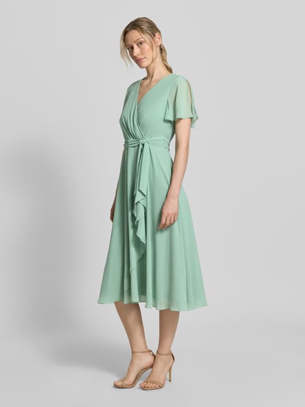 Frauenmodel trägt knielanges Cocktailkleid mit Bindegürtel von Christian Berg Cocktail aus Mintgrün-Chiffon, Taillierter Schnitt, Wickel-Optik; Ganzkörperansicht von vorne.