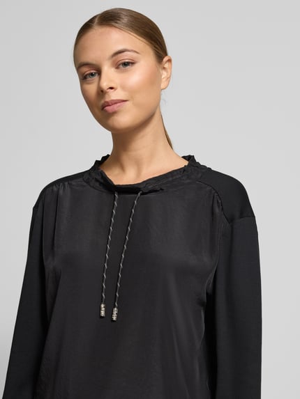 Frauenmodel trägt Longsleeve mit Rundhalsausschnitt von Marc Cain in Schwarz, glatte Textur, gerippte Ärmelabschlüsse, Nahaufnahme Oberkörper. Baumwoll-Elasthan-Mix, lockerer Schnitt, Tunnelzug am Halsausschnitt.