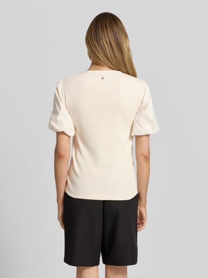 Vrouwenmodel draagt T-shirt met ballonmouwen model 'Jada' van MOS MOSH, katoenmix met stretch, fijnrib, V-hals, nauwsluitend model, ruglengte maat S: 60 cm, geheel in crèmekleur en zwart, gladde textuur, model met korte mouwen, detailrijke stofstructuur. Ganzkörperansicht Rückansicht.