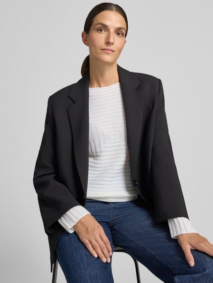 Frauenmodel trägt Strickpullover mit Strukturmuster von Emporio Armani in Weiß, kombiniert mit schwarzem Blazer und blauer Jeans, Nahaufnahme Oberkörper. Damen Strickpullover, taillierte Passform, Rundhalsausschnitt, langärmelig, Rückenlänge 35 cm, Ärmellänge 57 cm.