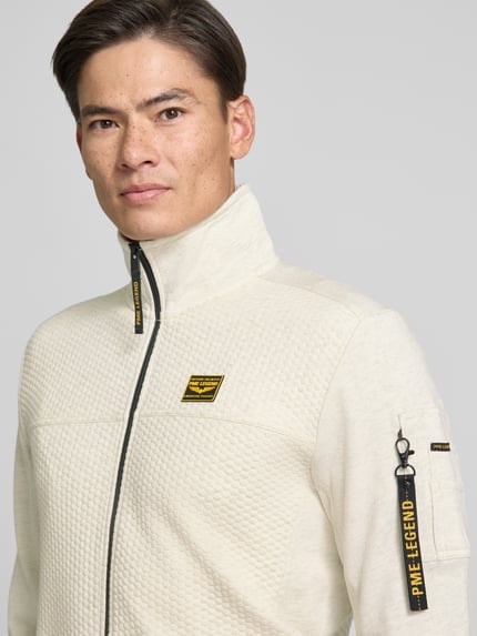 Männermodel trägt Sweatjacke mit Stehkragen und Label-Patch von PME Legend in Beige, Strukturmuster, Nahaufnahme Oberkörper. Material-Mix mit Baumwoll-Anteil, Reißverschluss mit Label-Detail, Reißverschlusstasche am Ärmel, seitliche Eingrifftaschen.