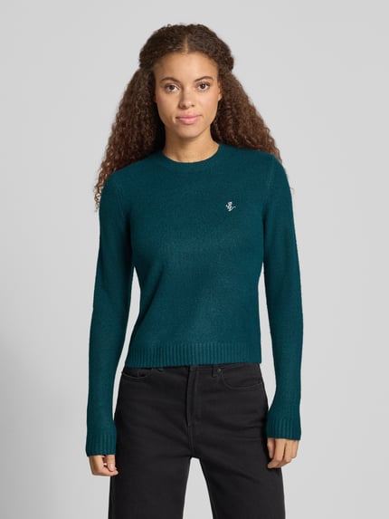 Kobiece modelka nosi Slim Fit Strickpullover aus elastischem Material-Mix von REVIEW, krój slim fit, elastyczna mieszanka materiałów, wyhaftowany motyw, prążkowane wykończenia, długość tyłu 51 cm, długość rękawów 61 cm, widoczne proste kolory i gładka tekstura, całkowity widok z przodu.