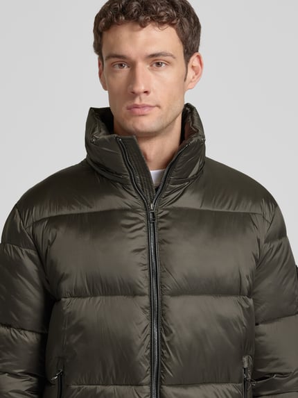 Männermodel trägt Steppjacke mit Reißverschlusstaschen van JOOP! Collection in olijfgroen met glanzende textuur, Nahaufnahme Oberkörper. Rechte snit, capuchon, rits, ritszakken, maat 50: ruglengte 75 cm, schouderbreedte 49 cm, borstbreedte 45 cm, mouwlengte 69 cm.