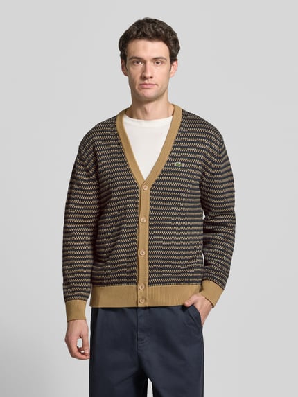 Männermodel trägt Cardigan mit Logo-Applikation von Lacoste, geometrisches Allover-Muster, Strickoptik, mehrfarbig, Ganzkörperansicht von vorne.