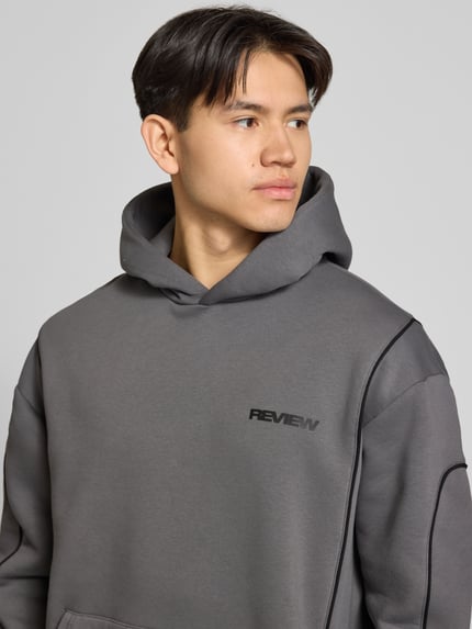 Männermodel trägt Hoodie mit Logo und Känguru-Tasche von REVIEW in grijs, regular fit, katoenmix, logo links op de borst, nahaufnahme oberkörper.