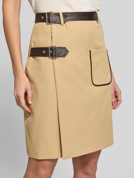 Frauenmodel trägt Midi-Rock mit Doppelgurt Modell 'SVEA' von BAUM & PFERDGARTEN in einfarbigem Beige, Wickeloptik, Lederoptik, Schnalle, Ziertasche, metalldetails, Webware, aufgesetzte Seitentasche, Nahaufnahme Oberkörper.
