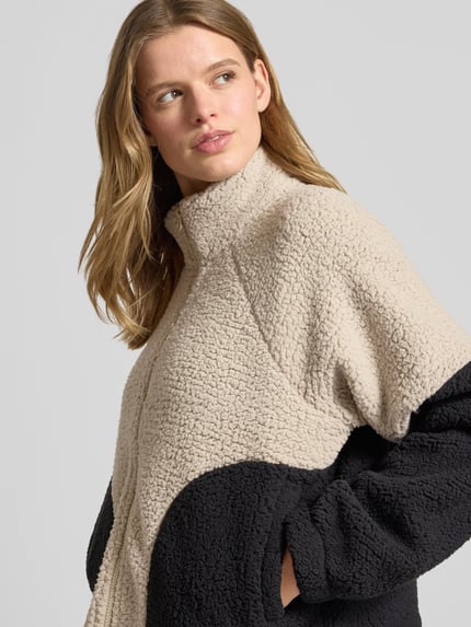 Frauenmodel trägt Fleecejacke mit Zweiwege-Reißverschluss Modell 'CUDDLE' von Noisy May, beige und schwarz, Relaxed-Fit, Stehkragen, Nahaufnahme Oberkörper, Rückenlänge 55 cm, Schulterbreite 50 cm, Brustweite 61 cm, Ärmellänge 60 cm.