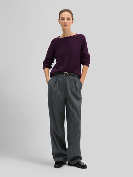 Frauenmodel trägt Regular Fit Pullover aus reiner Baumwolle von MARC O'POLO in unifarbener, violetter Strickoptik mit langen Ärmeln, U-Boot-Ausschnitt und seitlichen Eingrifftaschen, kombiniert mit weitem, grauem Stoffhose; Ganzkörperansicht von vorne.
