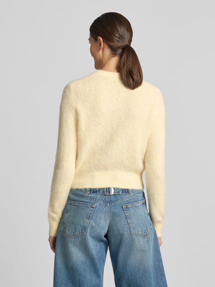 Frauenmodel trägt Strickjacke mit Rundhalsausschnitt Modell 'GLENNA' von DRYKORN in Creme, kombiniert mit blauer Jeans, Ganzkörperansicht Rückansicht. Regular Fit, feine Textur, Rückenlänge 52 cm, Ärmellänge 68 cm.