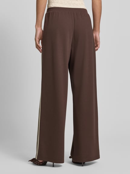 Frauenmodel trägt Jog Pants mit französischen Eingrifftaschen von Jake*s Casual in Braun, Wide-Leg-Fit, Material-Mix mit Stretch-Anteil, elastischer Bund, Ganzkörperansicht Rückansicht.