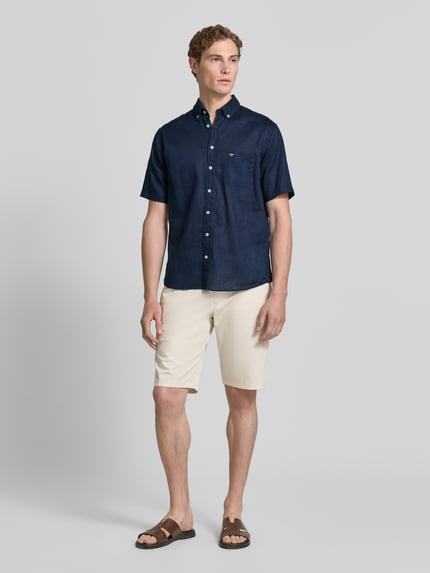 Männermodel trägt Slim Fit Freizeithemd aus Leinen-Baumwoll-Mix von Fynch-Hatton in Dunkelblau, Button-Down-Kragen, Brusttasche, Label-Stitching, Knopfleiste, Kombi mit heller Shorts, Ganzkörperansicht von vorne. Rückenlänge bei Größe M: 74 cm, Schulterbreite: 48 cm, Ärmellänge: 26 cm.