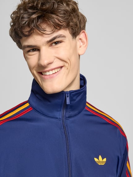 Männermodel trägt Regular Fit Sweatjacke mit Logo-Stitching Modell 'FIREBIRD TT' von adidas Originals, blauw met gekleurde strepen, nahaufnahme oberkörper.