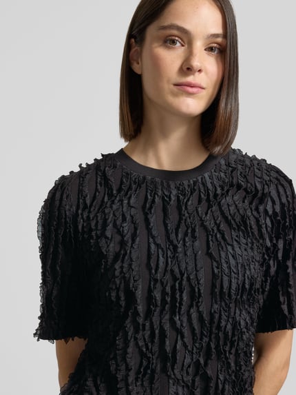 Frauenmodel trägt Regular Fit T-Shirt mit Rüschenbesatz und Rundhalsausschnitt von Jake*s Collection, Material-Mix mit Stretch-Anteil, schwarze Farbe, feine Rüschentextur, Nahaufnahme Oberkörper.