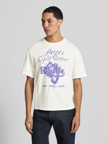 Männermodel trägt Regular Fit T-Shirt aus reiner Viskose von RHUDE mit Label-Print, helle Textur, Rundhalsausschnitt, Ganzkörperansicht von vorne.