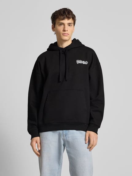 Männermodel trägt Relaxed Fit Hoodie aus reiner Baumwolle Modell 'NYROHOOD' von Hugo Blue in Schwarz mit Label Print und Kängurutasche, Ganzkörperansicht von vorne.