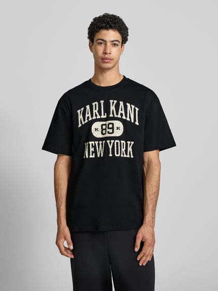 Männermodel trägt Oversized Fit T-Shirt mit Logo-Print Modell 'Heritage' von KARL KANI in Schwarz aus Reine Baumwolle, Rundhalsausschnitt, Rückenlänge bei Größe M 71 cm, Herren T-Shirt, Ganzkörperansicht von vorne.
