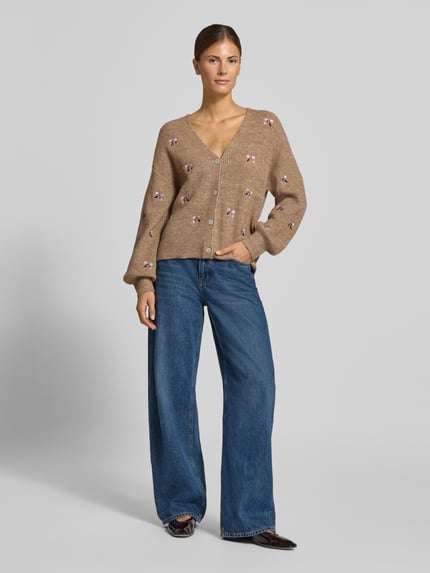 Frauenmodel trägt Relaxed Fit Strickjacke aus elastischem Woll-Mix Modell 'FLEUR' von YAS, beige mit floraler Stickerei, V-Ausschnitt, Knopfleiste, kombiniert mit blauer weiter Jeans, Ganzkörperansicht von vorne.