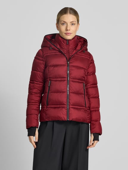 Frauenmodel trägt Steppjacke mit Kapuze und Reißverschlusstaschen von MICHAEL Michael Kors in Weinrot, sichtbare Steppnähte, glatte Textur, Ganzkörperansicht von vorne.