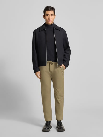 Männermodel trägt Stoffhose mit elastischem Bund von MCNEAL in Beige, Material-Mix mit Viskose-Anteil, lockerer Schnitt, Tunnelzug, Knopf- und Reißverschluss, französische Eingrifftaschen, Gesäßtaschen, Ganzkörperansicht von vorne.
