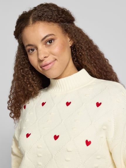 Modelka nosi Regular Fit Strickpullover mit Stitchings von NOISY MAY®, kremowa dzianina z czerwonymi serduszkami, krój regular fit, stebnowania, stójka, model 'MILLIE', zbliżenie na górną część ciała.