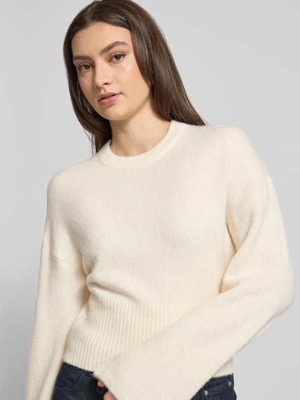Vrouwenmodel draagt gebreide pullover met ronde hals, wijde mouwen, regular fit in materiaalmix met alpacawol, model 'Stella' van Gina Tricot, ruglengte maat S 55 cm, mouwlengte maat S 60 cm, lichte kleur, zachte textuur, Nahaufnahme bovenlichaam.