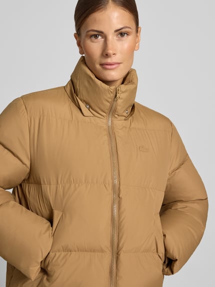 Frauenmodel trägt beige Pufferjacke mit Kapuze von LACOSTE, unifarbene Optik, Logo-Stitching, Reißverschluss, Nahaufnahme Oberkörper.
