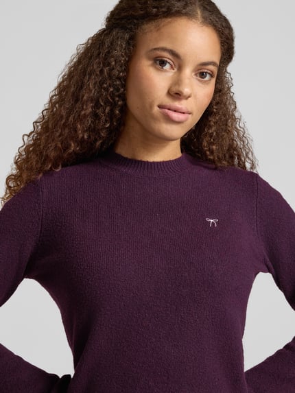 Modelka kobieca ma na sobie Slim Fit Strickpullover aus elastischem Material-Mix od REVIEW, elastyczna mieszanka materiałów, krój slim fit, wyhaftowany motyw, prążkowane wykończenia, długość tyłu w rozmiarze S: 51 cm, długość rękawów w rozmiarze S: 61 cm, fiolet, faktura dzianiny, prosty fason, widoczna struktura materiału, zbliżenie na górną część ciała.