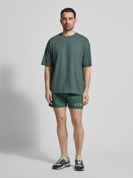 Męski model nosi szorty z elastycznym pasem od HALO, mieszanka materiałów z dodatkiem streczu, krój regular fit, nadruk z logo, szerokość w rozmiarze M: 40 cm, długość wewnętrzna nogawki w rozmiarze M: 12 cm, zielony kolor, gładka faktura, widok całej sylwetki z przodu.