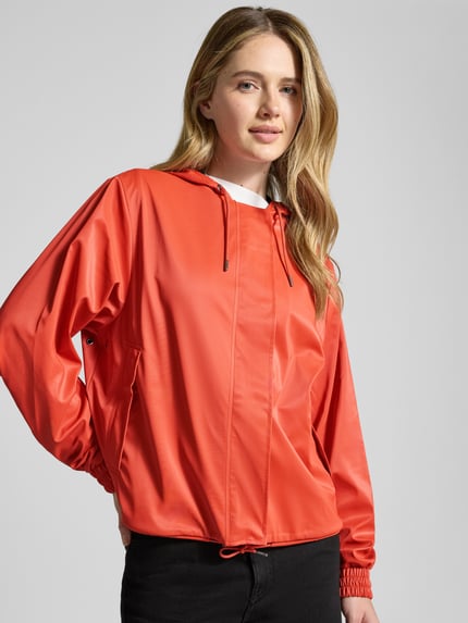 Modelka nosi Jacke mit Abdeckleiste od RAINS w kolorze czerwonym, prosta forma, kaptur ze ściągaczem, długi zamek z listwą, ściągacz w talii, elastyczne mankiety, długość pleców w rozmiarze S 58 cm, szerokość ramion w rozmiarze S 43 cm, szerokość w klatce piersiowej w rozmiarze S 55 cm, długość rękawa w rozmiarze S 63 cm, matowa faktura, widok z przodu, zbliżenie na górną część ciała.