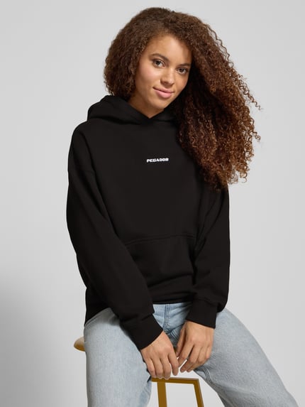 Frauenmodel trägt Oversized Hoodie mit Label-Stitching Modell 'CLARITA' von Pegador, Baumwoll-Mix, schwarze Farbe, Nahaufnahme Oberkörper.